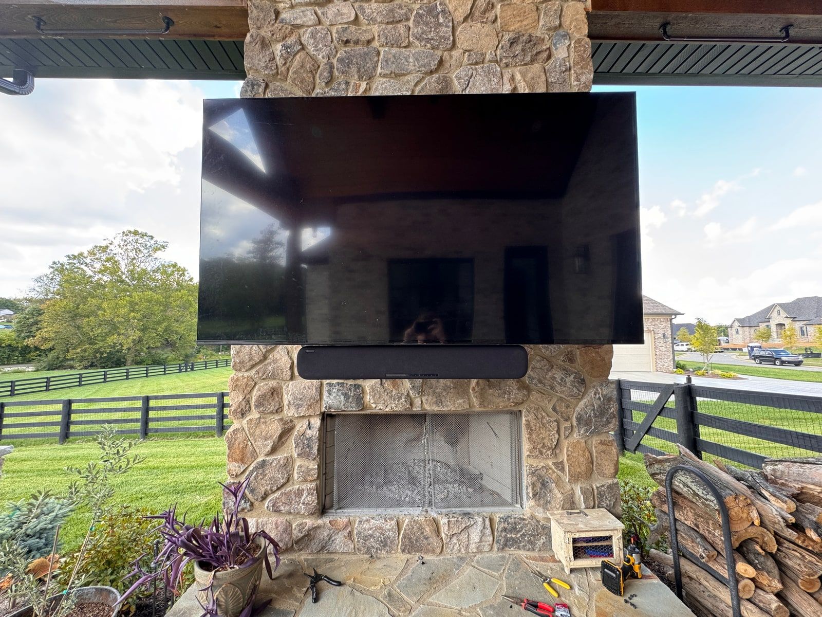 A weatherproof television mounted on an outdoor stone fireplace under a covered patio, facing a fenced yard.